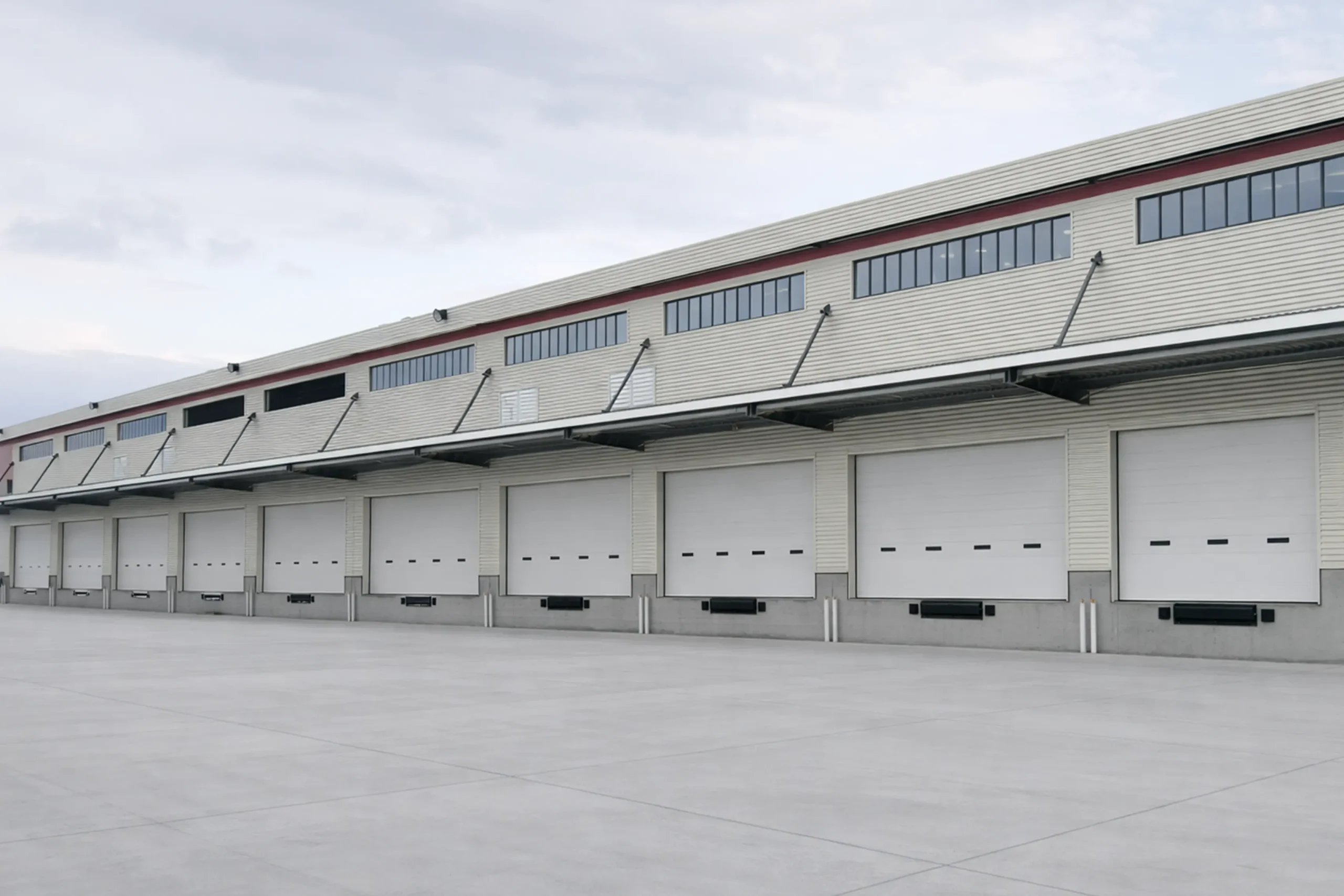Exterior view of a large modern distribution center featuring a continuous row of white automatic industrial sectional doors, providing high-speed access for logistics docks.